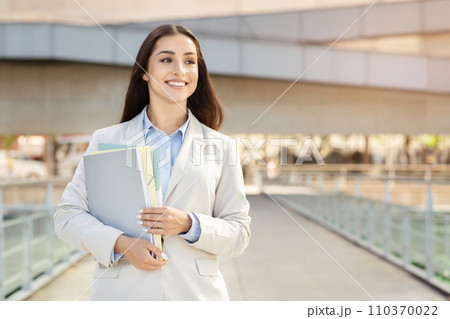A contented young woman secretary, manager in a smart suit holds files, walking with confidence 110370022