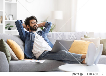 Happy indian man in casual wear reclining on sofa with his laptop Happy indian man in casual wear reclining on sofa with his laptop 110370307