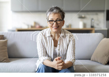 Smiling elderly woman sitting on couch in living room interior Smiling elderly woman sitting on couch in living room interior 110371258