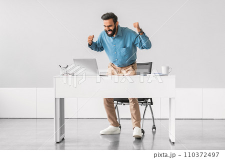 Energetic indian businessman celebrating victory at his desk. Energetic indian businessman celebrating victory at his desk. 110372497