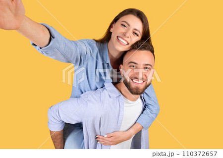 Happy loving european couple taking selfie together, cheerful young woman piggybacking her boyfriend, yellow background Happy loving european couple taking selfie together, cheerful young woman piggybacking her boyfriend, yellow background 110372657