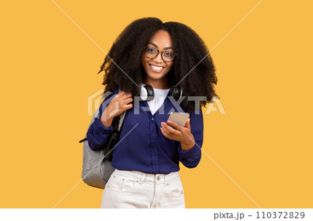 Excited black lady student in glasses with backpack, using cellphone and smiling, on yellow background Excited black lady student in glasses with backpack, using cellphone and smiling, on yellow background 110372829