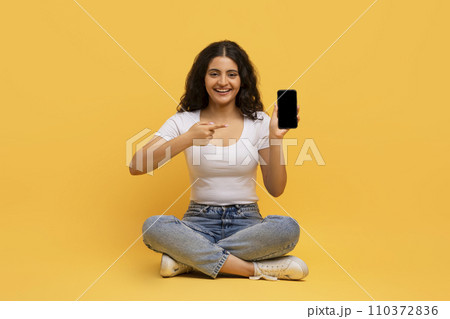 Cheerful attractive young hindu lady with smartphone sitting on floor Cheerful attractive young hindu lady with smartphone sitting on floor 110372836