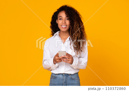 Happy african american woman holds her mobile phone, yellow background 110372860