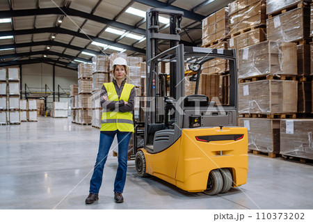 Female forklift driver standing by forklift. Warehouse worker preparing products for shipmennt, delivery, checking stock in warehouse. 110373202