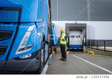 Warehouse receiver standing by blue truck, holding tablet, looking at cargo details, checking delivered items, goods against order. Warehouse receiver standing by blue truck, holding tablet, looking at cargo details, checking delivered items, goods against order. 110373346