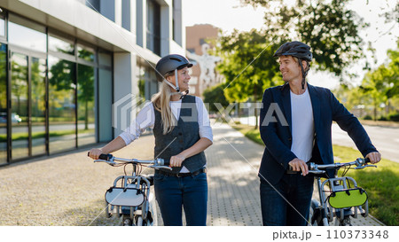 Spouses commuting through the city, talking and walking by bike on street. Middle-aged city commuters traveling from work by bike after a long workday. Spouses commuting through the city, talking and walking by bike on street. Middle-aged city commuters traveling from work by bike after a long workday. 110373348