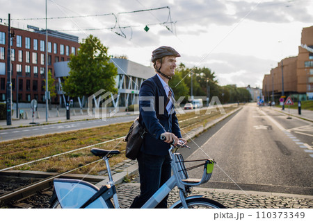 Handsome middle-aged man commuting through the city by bike, walking next to bike crossing the road. Male city commuter traveling from work by bike after a long workday. Handsome middle-aged man commuting through the city by bike, walking next to bike crossing the road. Male city commuter traveling from work by bike after a long workday. 110373349