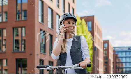 Beautiful middle-aged woman commuting through the city by bike, phone calling on smartphone. Female city commuter traveling from work by bike after long workday. 110373363