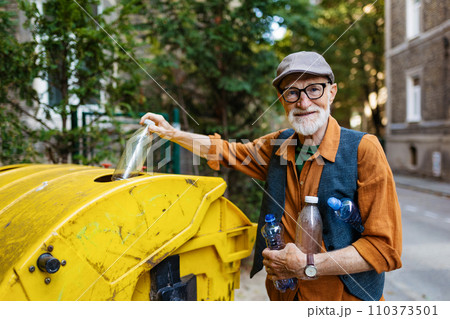 Senior man throwing plastic waste, bottles into recycling container in front apartment. Elderly man sorting the waste according to material into colored bins. Senior man throwing plastic waste, bottles into recycling container in front apartment. Elderly man sorting the waste according to material into colored bins. 110373501