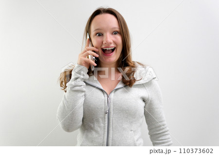 Image of beautiful smiling blonde woman talking on cellphone and laughing, having business call on mobile phone, standing in red blazer against gray background. Business and technology 110373602