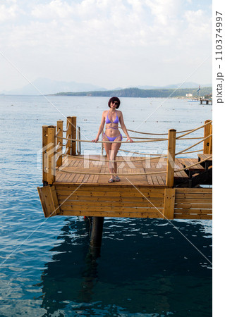 A girl in a bikini sunbathes while standing on the edge of a wooden pier A girl in a bikini sunbathes while standing on the edge of a wooden pier 110374997