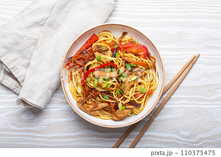 White bowl with Chow Mein or Lo Mein, traditional Chinese stir fry noodles with meat and vegetables, served with chopsticks top view on rustic white wooden background table 110375475