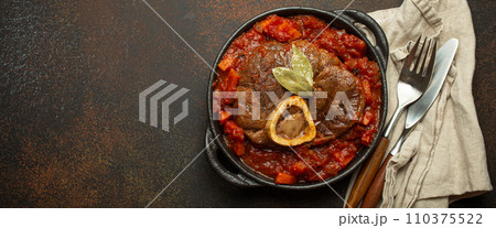 Traditional Italian dish Ossobuco all Milanese made with cut veal shank meat with vegetable tomato sauce served in black casserole pan top view on rustic brown background 110375522