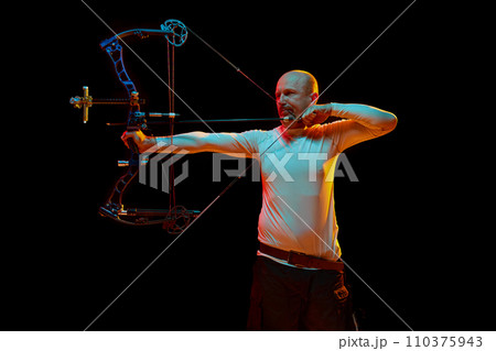 Man, professional archer training, aiming archery bow into target against black studio background in neon light. Archery sport, competition Man, professional archer training, aiming archery bow into target against black studio background in neon light. Archery sport, competition 110375943