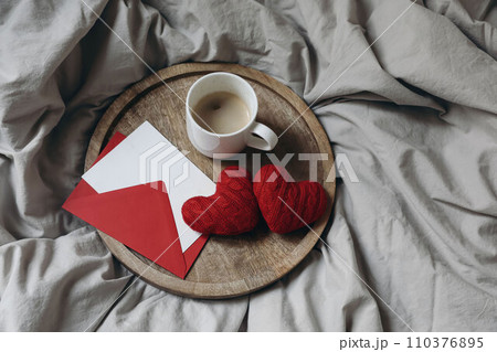 Valentines day still life. Blank greeting card, invitation mockup, red envelope on wooden tray. Couple of red textile hearts. Cup of coffee in bed. Linen duvet cover. Love concept. Flatlay, top view. 110376895