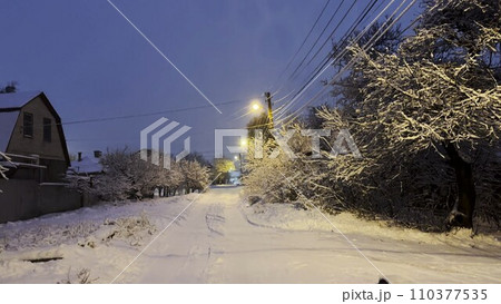 Snowy country road at evening time. Street lamp illuminating snow trail at winter night. Beautiful nature landscape with falling snowflakes at background. 110377535