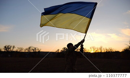 Male ukrainian army soldier going and waving flag of Ukraine on meadow at dusk. Young man in camouflage uniform walking with raised blue-yellow banner as symbol of victory against russian aggression Male ukrainian army soldier going and waving flag of Ukraine on meadow at dusk. Young man in camouflage uniform walking with raised blue-yellow banner as symbol of victory against russian aggression 110377777