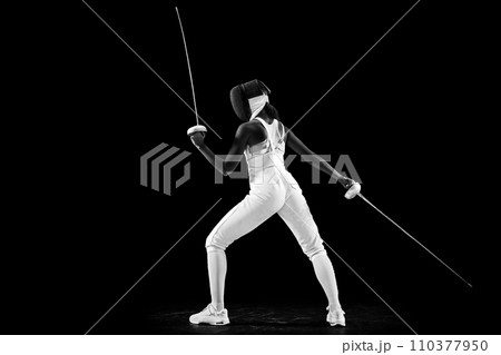 Striking profile of two-sword-wielding sportswoman, radiating power and grace, athleticism and dedication against black studio background. 110377950