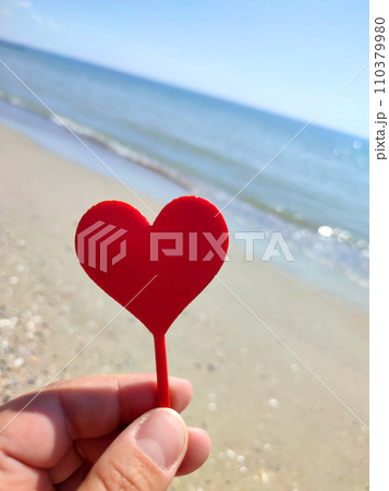 Person holding in fingers hand stick in shape red heart on background sea and sea waves, blue sky in seashore on sunny summer day close-up. Concept love romance amour St Valentines Day 110379980
