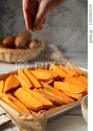 Cooking sweet potato wedges, fries on a baking pan. Hand adding herbs and spices. Cooking sweet potato wedges, fries on a baking pan. Hand adding herbs and spices. 110380529