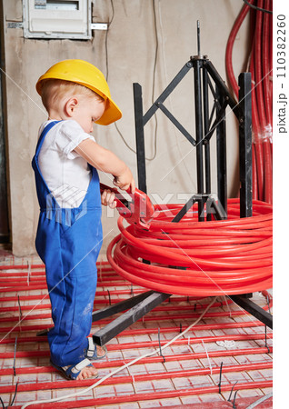 Full length of child in work overalls cutting pipe with special tool at home. Kid in construction helmet using pipe cutter while installing underfloor heating system in apartment under renovation. 110382260