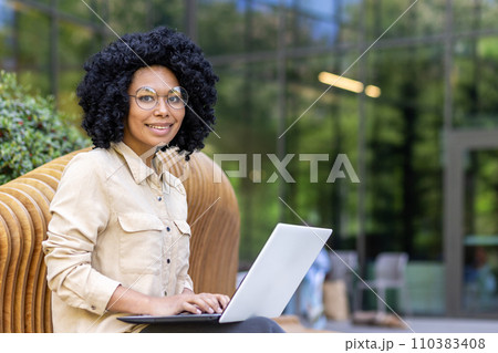 Portrait of an African American young female student sitting on a bench outside a university campus studying online on a laptop. Smiling and looking at the camera. Portrait of an African American young female student sitting on a bench outside a university campus studying online on a laptop. Smiling and looking at the camera. 110383408