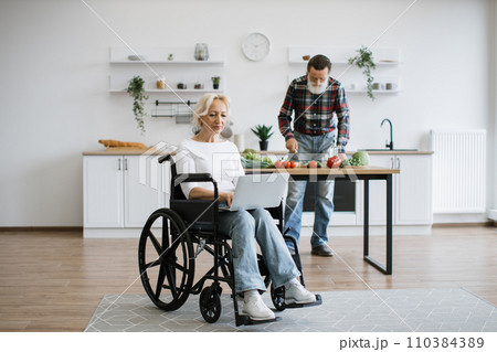 Old woman sits in wheelchair and works on laptop while her husband prepares healthy salad Old woman sits in wheelchair and works on laptop while her husband prepares healthy salad 110384389