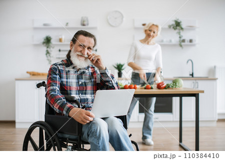 Old man sits in wheelchair and works on laptop while his wife prepares healthy salad. Old man sits in wheelchair and works on laptop while his wife prepares healthy salad. 110384410