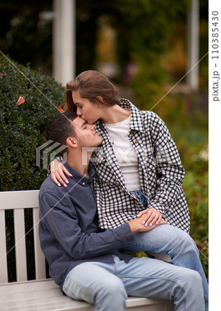 Autumn Romance: Young Couple Embracing and Kissing on Park Bench. Young couple kissing and rejoices at the lake. lovely young couple kissing outdoors in autumn. Embraced in Autumn's Warmth: Couple's 110385430