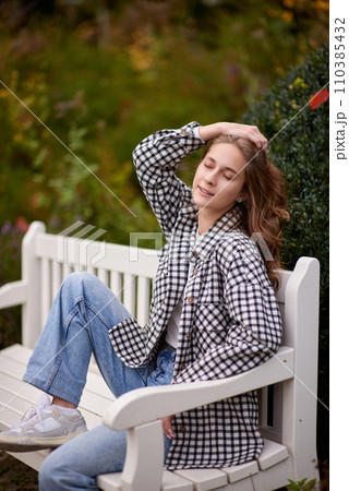 Beautiful teen blonde girl sits on a bench in an autumn park . attractive young smiling girl sitting on a bench in an autumn park 110385432