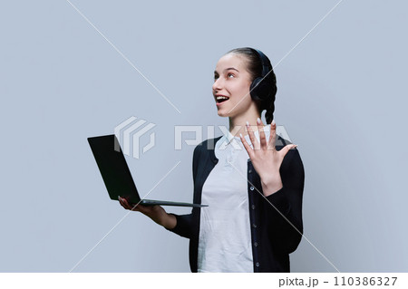 Teen girl student wearing headphones using laptop, on grey background Teen girl student wearing headphones using laptop, on grey background 110386327