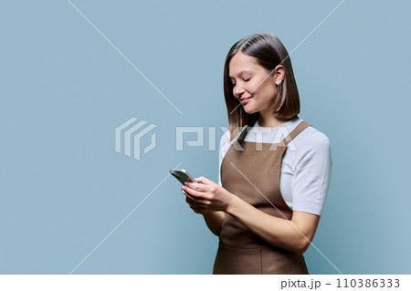 Portrait of young woman in apron holding smartphone in hands on blue background 110386333