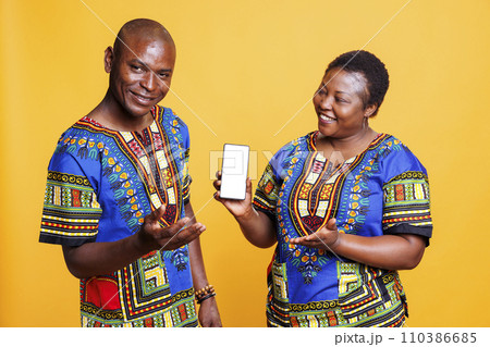 Smiling black couple wearing ethnic closes showing smartphone with white blank screen mockup. Cheerful man and woman presenting mobile phone with empty touchscreen for app advertising 110386685