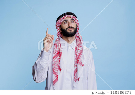 Muslim man dressed in traditional clothes pointing upwards with finger. Young arab wearing islamic thobe and checkered headdress showing and looking up while posing in studio 110386875