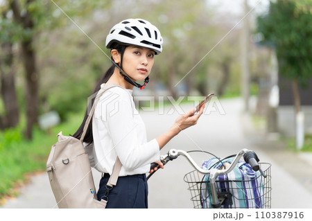 サイクリングヘルメットを被ってスマホ地図を見て周囲を確認しながら自転車を引いて歩く若い女性 サイクリングヘルメットを被ってスマホ地図を見て周囲を確認しながら自転車を引いて歩く若い女性 110387916