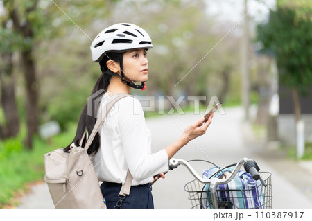サイクリングヘルメットを被ってスマホ地図を見て周囲を確認しながら自転車を引いて歩く若い女性 サイクリングヘルメットを被ってスマホ地図を見て周囲を確認しながら自転車を引いて歩く若い女性 110387917
