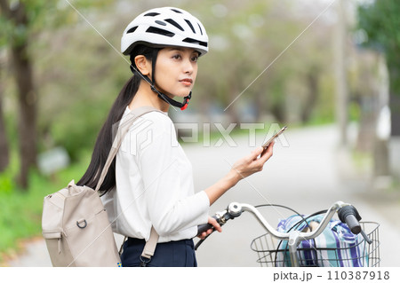 サイクリングヘルメットを被ってスマホ地図を見て周囲を確認しながら自転車を引いて歩く若い女性 サイクリングヘルメットを被ってスマホ地図を見て周囲を確認しながら自転車を引いて歩く若い女性 110387918