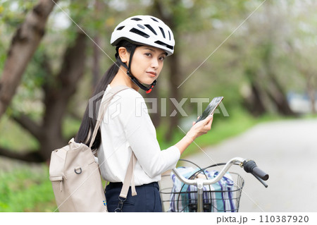 サイクリングヘルメットを被ってスマホ地図を見て周囲を確認しながら自転車を引いて歩く若い女性 サイクリングヘルメットを被ってスマホ地図を見て周囲を確認しながら自転車を引いて歩く若い女性 110387920