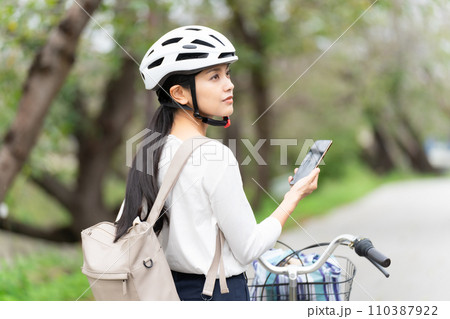 サイクリングヘルメットを被ってスマホ地図を見て周囲を確認しながら自転車を引いて歩く若い女性 110387922