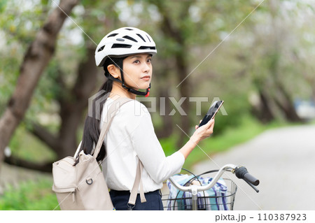 サイクリングヘルメットを被ってスマホ地図を見て周囲を確認しながら自転車を引いて歩く若い女性 サイクリングヘルメットを被ってスマホ地図を見て周囲を確認しながら自転車を引いて歩く若い女性 110387923