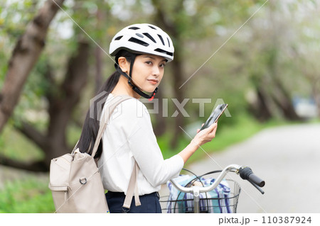 サイクリングヘルメットを被ってスマホ地図を見て周囲を確認しながら自転車を引いて歩く若い女性 サイクリングヘルメットを被ってスマホ地図を見て周囲を確認しながら自転車を引いて歩く若い女性 110387924