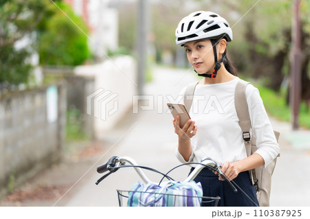 サイクリングヘルメットを被ってスマホ地図を見て周囲を確認しながら自転車を引いて歩く若い女性 サイクリングヘルメットを被ってスマホ地図を見て周囲を確認しながら自転車を引いて歩く若い女性 110387925