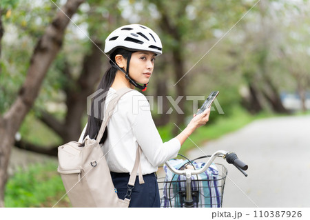 サイクリングヘルメットを被ってスマホ地図を見て周囲を確認しながら自転車を引いて歩く若い女性 サイクリングヘルメットを被ってスマホ地図を見て周囲を確認しながら自転車を引いて歩く若い女性 110387926