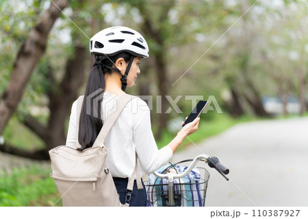 サイクリングヘルメットを被ってスマホ地図を見て周囲を確認しながら自転車を引いて歩く若い女性 サイクリングヘルメットを被ってスマホ地図を見て周囲を確認しながら自転車を引いて歩く若い女性 110387927