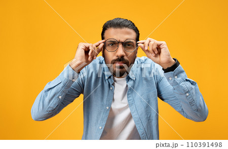 Focused man in a blue shirt adjusting his glasses, looking seriously at the camera 110391498