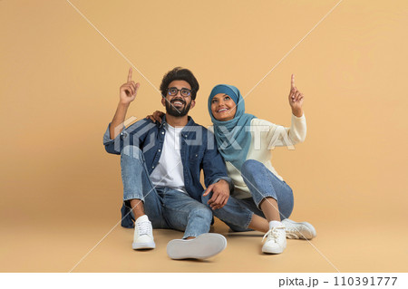 Joyful muslim couple sitting together against beige background and pointing upwards Joyful muslim couple sitting together against beige background and pointing upwards 110391777