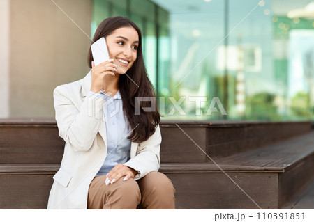 Smiling businesswoman in a light blazer talking on a smartphone while sitting on an outdoor bench 110391851