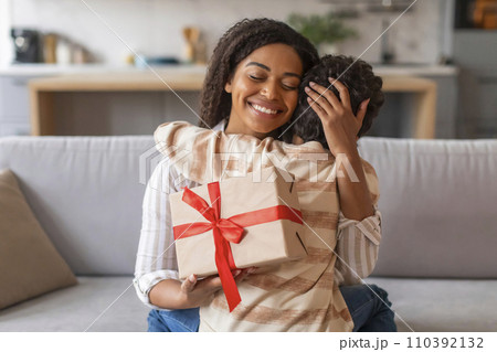 Joyful black mother holding wrapped gift box and affectionately embracing her son Joyful black mother holding wrapped gift box and affectionately embracing her son 110392132
