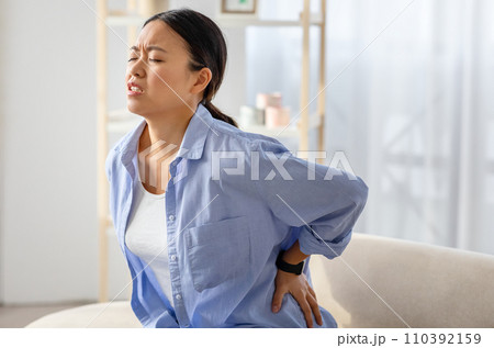 Chinese woman rubbing lower back, sitting on couch at home Chinese woman rubbing lower back, sitting on couch at home 110392159